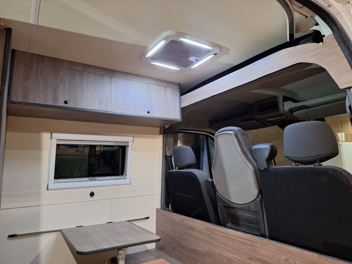 Menjador de la furgoneta camper amb taula abatible, finestra amb cortina i claraboia amb llum LED